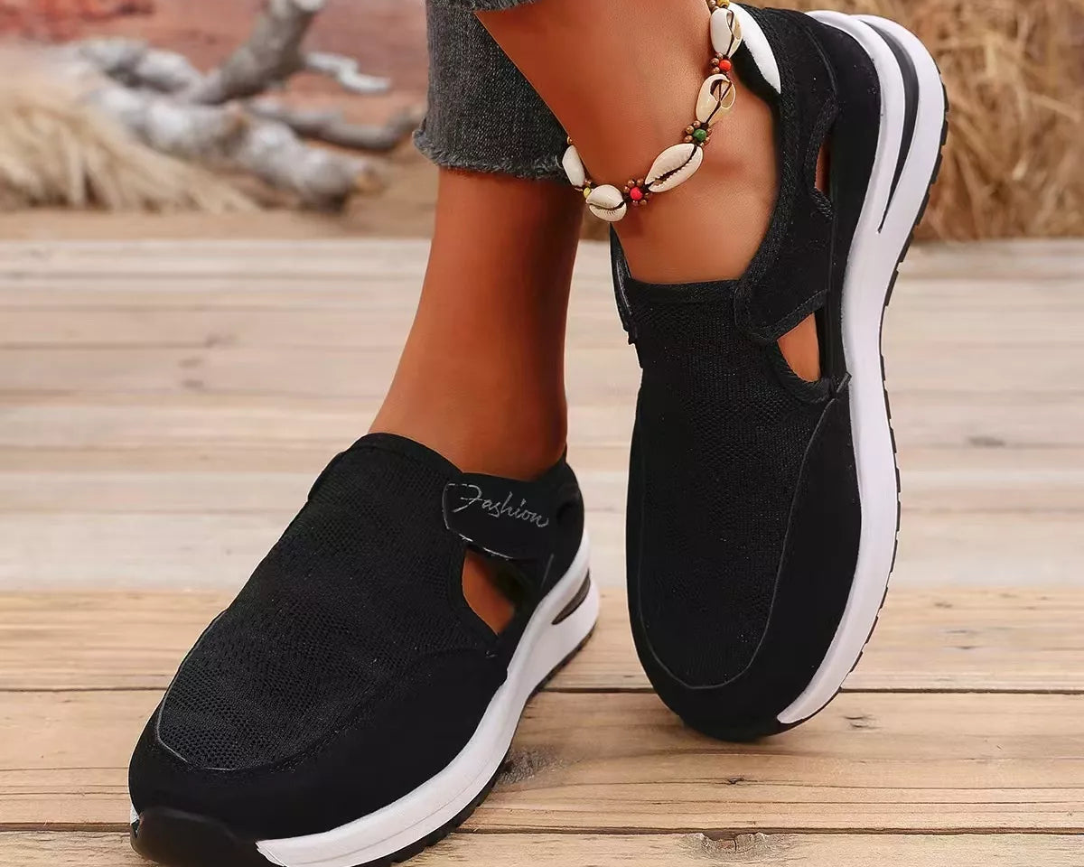 NEWYORKLISTED SHOE COLLECTION: A person wearing black slip-on platform sneakers with white soles, standing on a wooden deck. The shoes have cutout details and a textured fabric upper.
