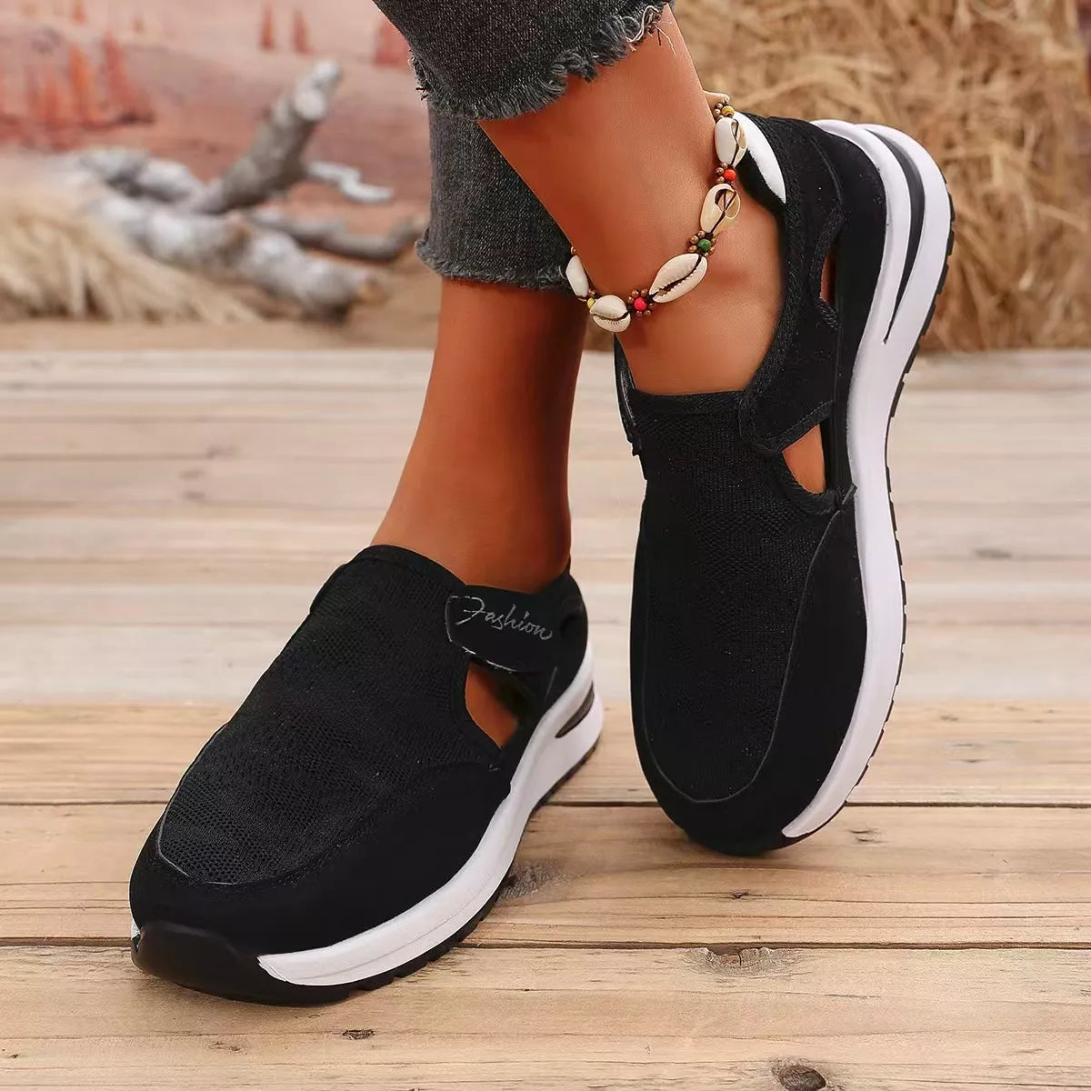 NEWYORKLISTED SHOE COLLECTION: A person wearing black slip-on platform sneakers with white soles, standing on a wooden deck. The shoes have cutout details and a textured fabric upper.