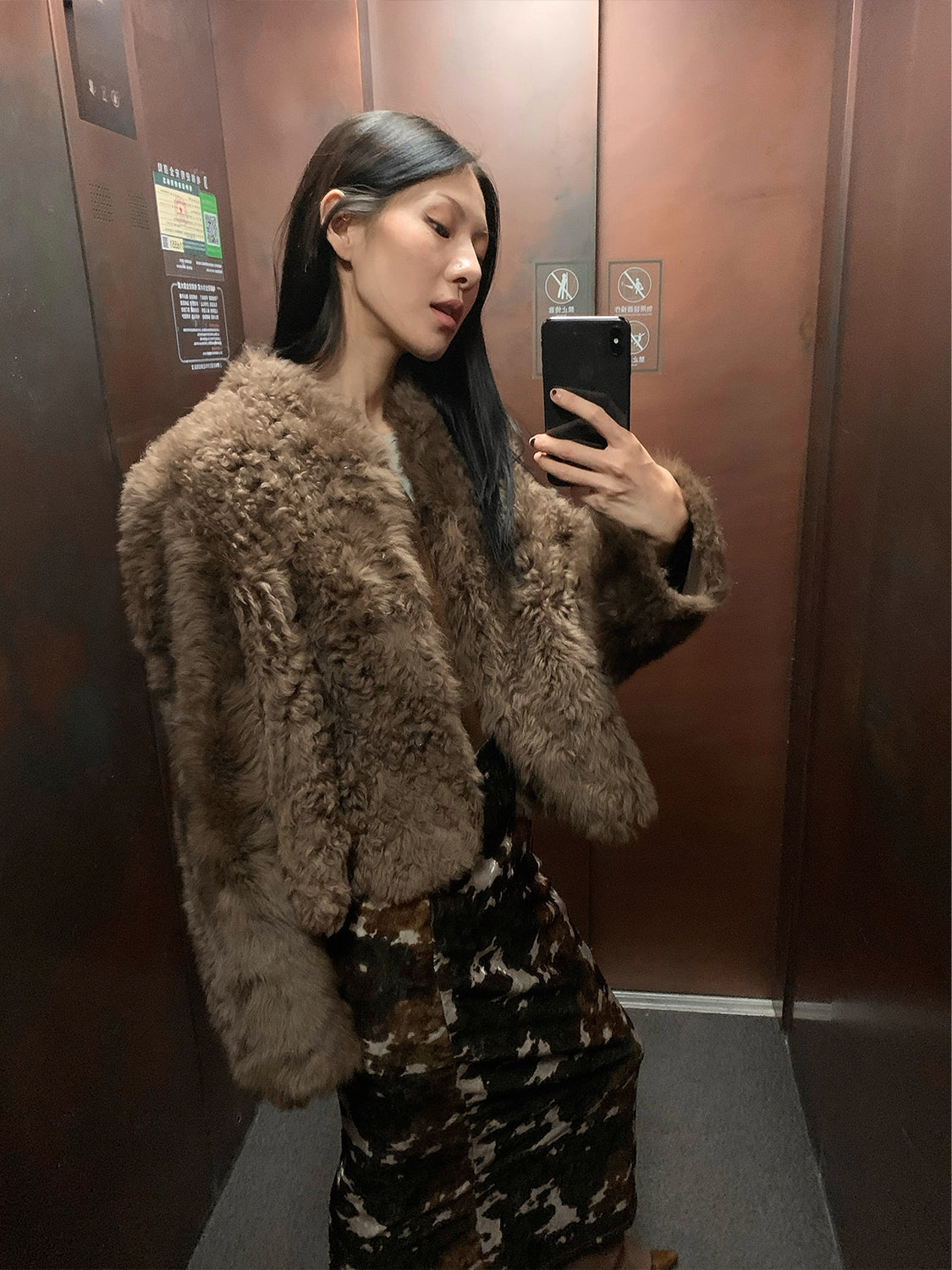 NEWYORKLISTED STYLE COLLECTION: Person wearing a fluffy brown coat taking a mirror selfie in an elevator.