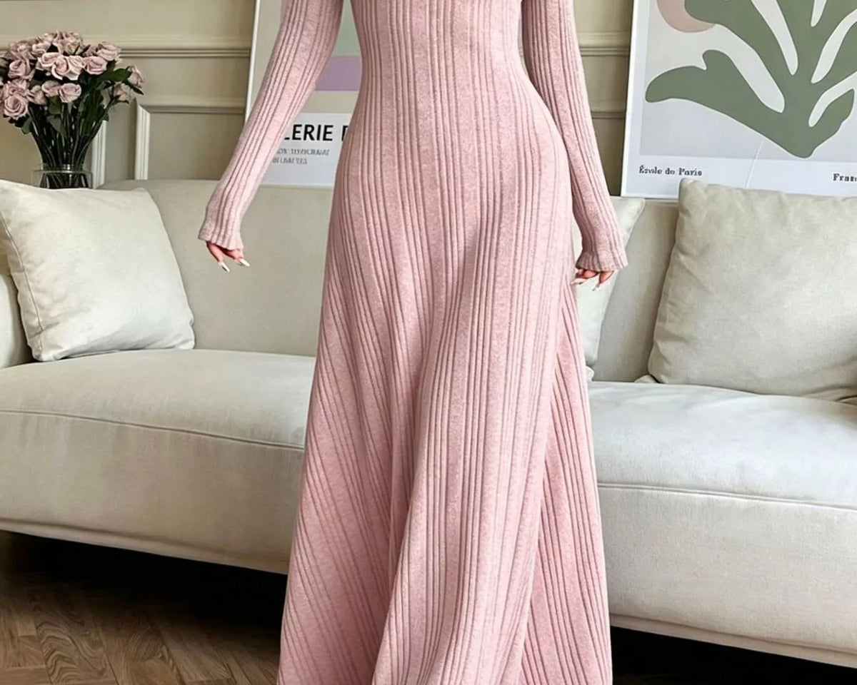 NEWYORKLISTED STYLE COLLECTION: A woman wearing a long-sleeve, floor-length pink ribbed knit dress, standing in a living room setting with a beige sofa and framed wall art in the background.