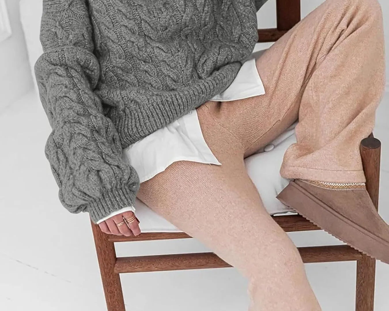 NEWYORKLISTED STYLE COLLECTION: Model wearing a gray cable knit sweater and beige pants sitting on a wooden chair.