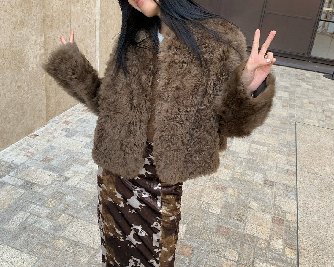Person wearing a brown fur coat and patterned pants standing on a sidewalk.