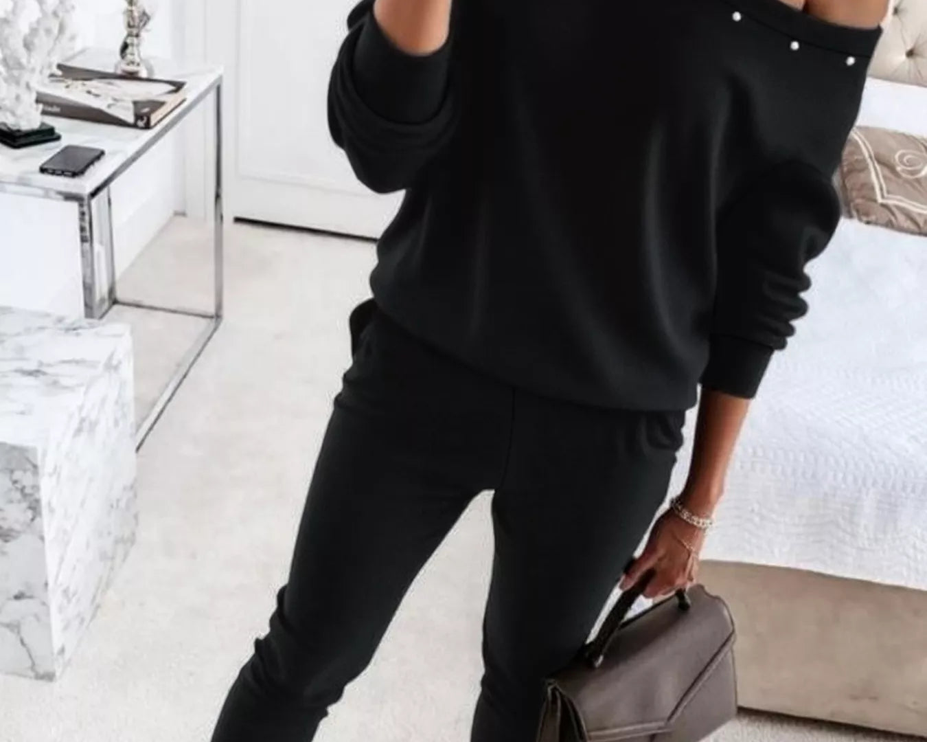 NEWYORKLISTED STYLE COLLECTION: Person wearing a black off-shoulder top with pearl details and black jogger pants, holding a brown handbag in a modern living room.