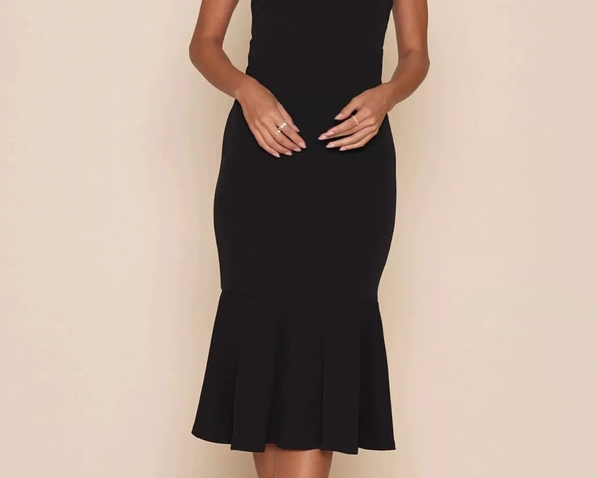 NEWYORKLISTED STYLE COLLECTION: A woman modeling a sleeveless black midi dress with a square neckline, wide shoulder straps, and a flared ruffled hem against a plain beige studio background.