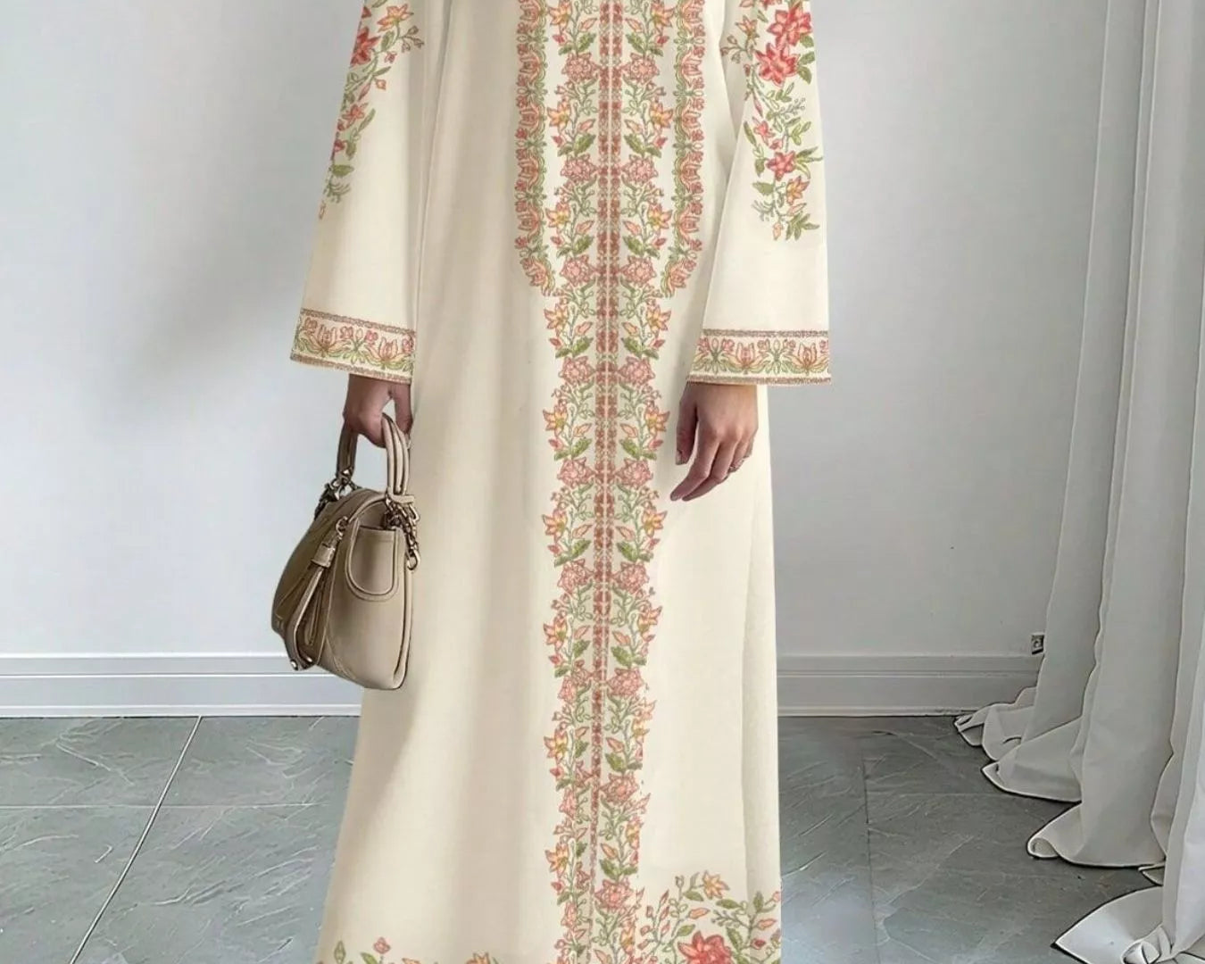 NEWYORKLISTED STYLE COLLECTION: Woman wearing a long, cream-colored dress with floral embroidery in a room with light gray flooring and white walls.