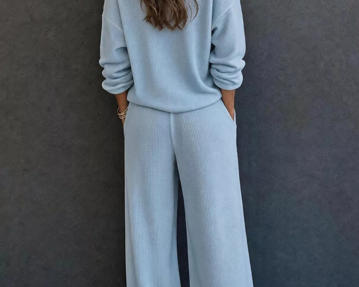 NEWYORKLISTED STYLE & COMFORT COLLECTION: A woman viewed from behind wearing a light blue knit matching set consisting of a long-sleeve top and wide-leg pants, standing against a dark gray textured wall.