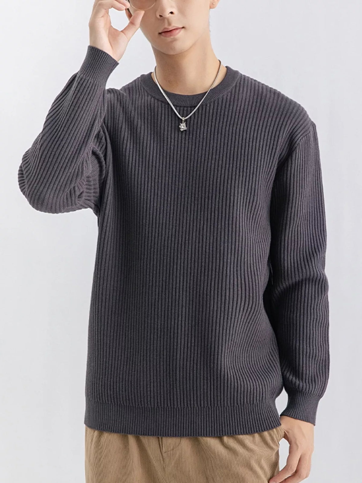 NEWYORKLISTED ATLAS MEN'S COLLECTION: A man wearing a dark gray ribbed knit crewneck sweater, paired with tan pants and a silver necklace, against a plain light gray background.