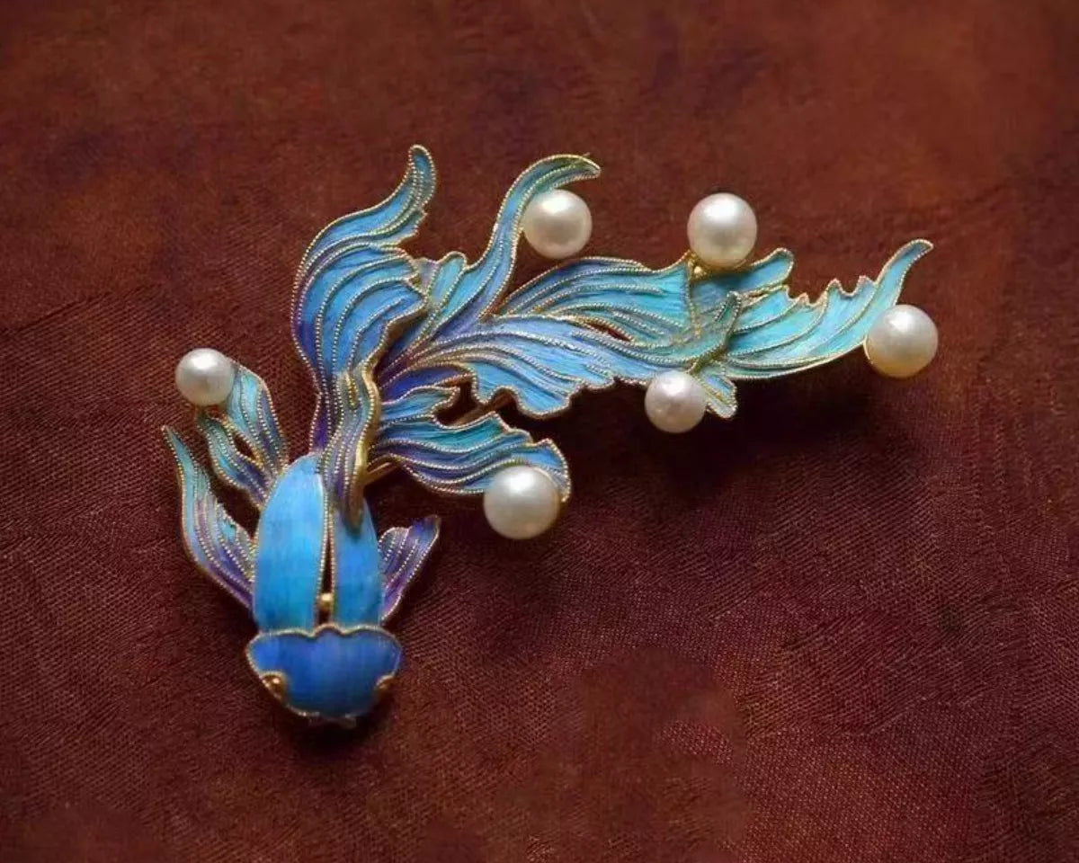 NYLD GLAMOUR By NEWYORKLISTED: Jewelry brooch with blue fish design and pearl-like beads on a brown fabric background
