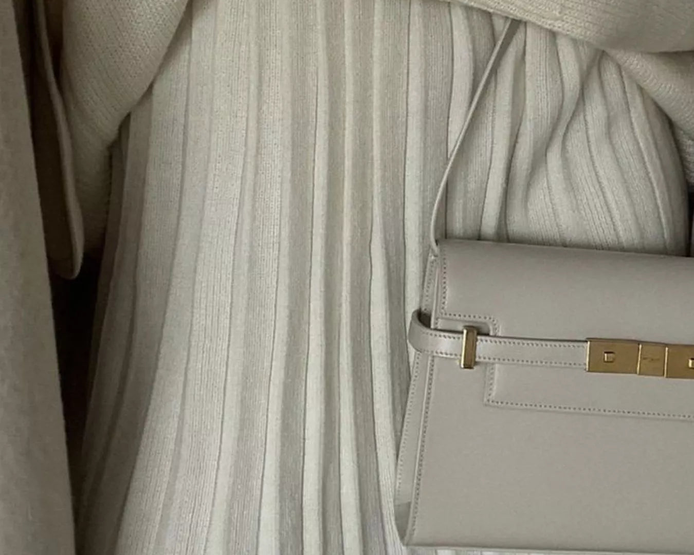 Person wearing a beige sweater and pleated skirt holding a matching handbag.