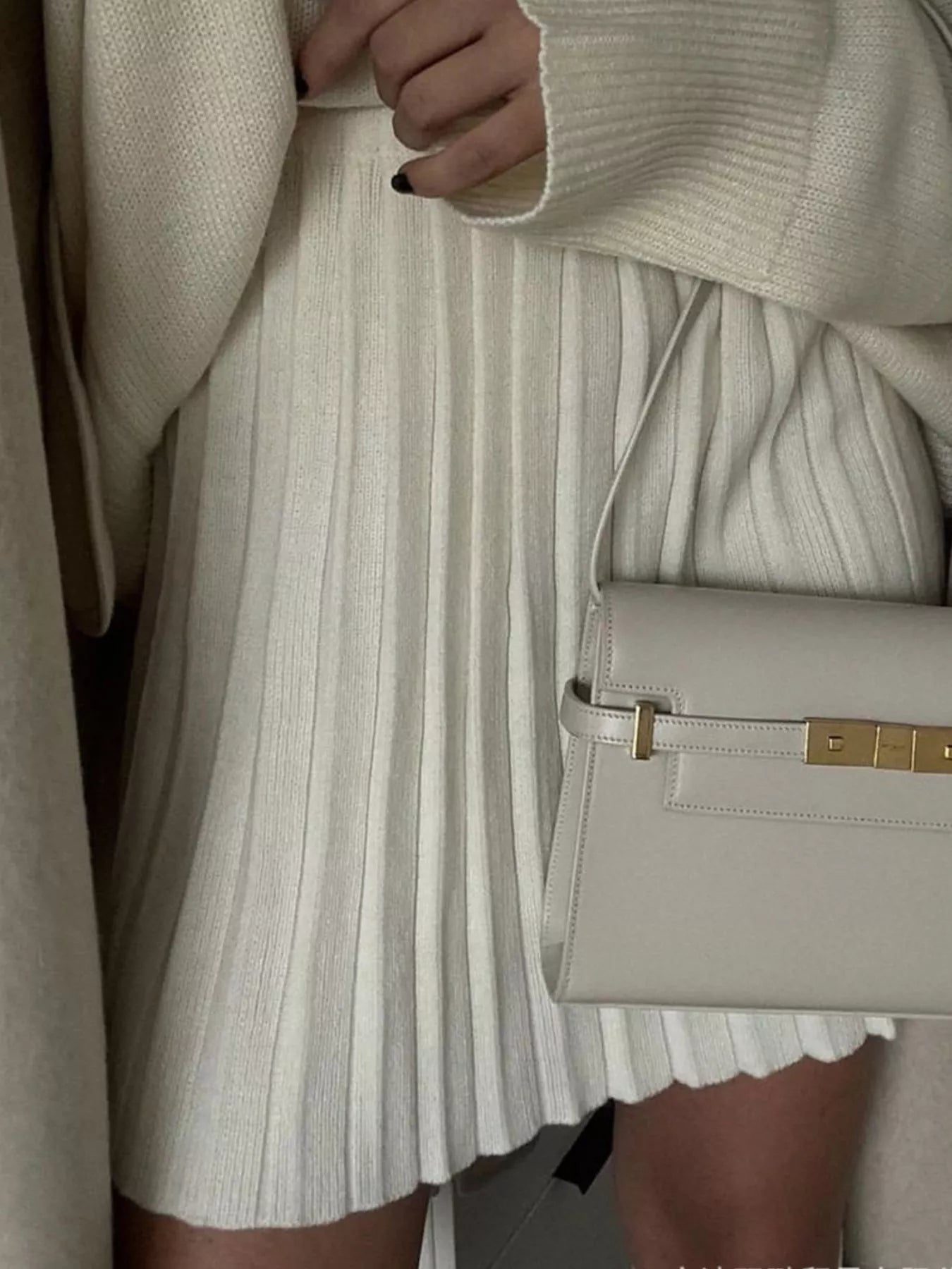 Person wearing a beige sweater and pleated skirt holding a matching handbag.