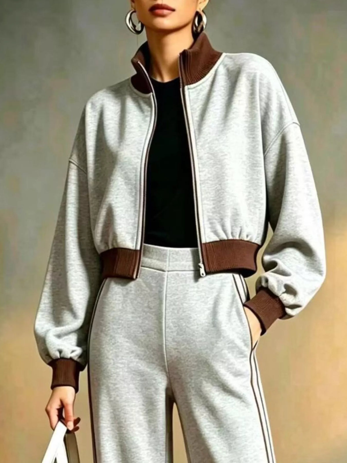 NEWYORKLISTED STYLE COLLECTION: A person wearing a light gray cropped zip-up jacket with brown ribbed collar, hem, and cuffs, paired with matching light gray sweatpants and a black top, against a soft neutral studio background.