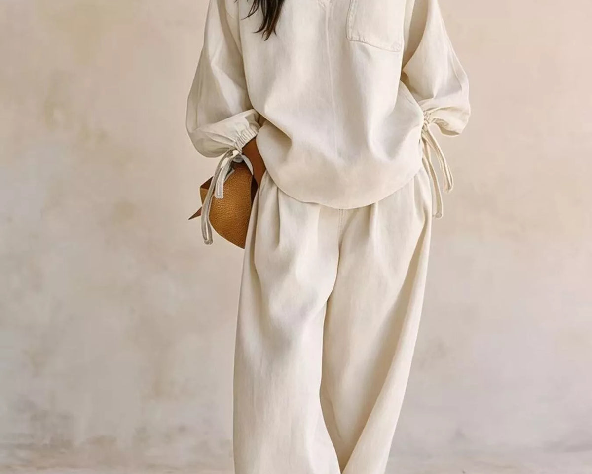 NEWYORKLISTED STYLE COLLECTION: A woman modeling a matching cream-colored lounge set consisting of a long-sleeve V-neck top with tie details at the cuffs and matching wide-leg trousers, set against a textured beige wall.