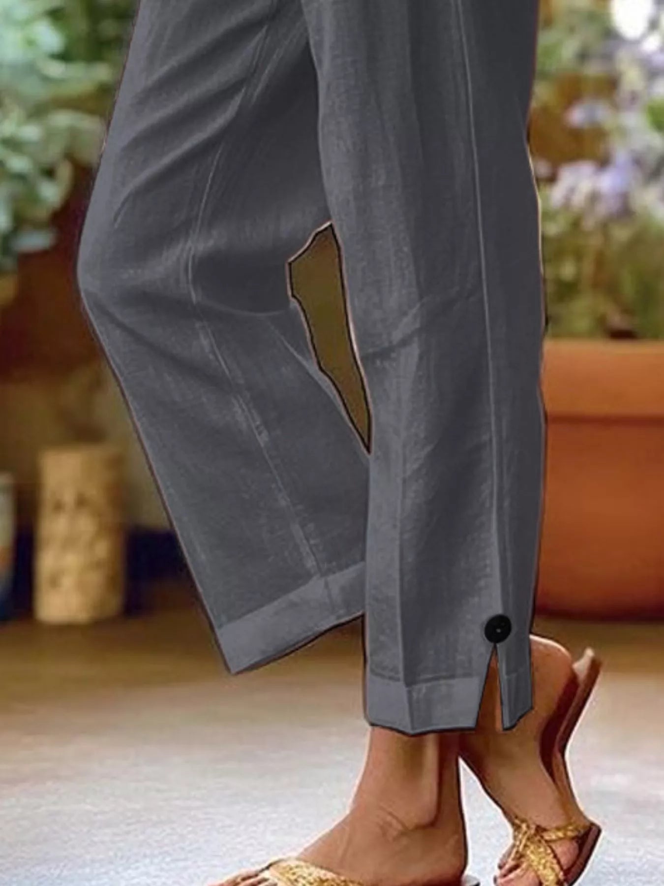 NEWYORKLISTED STYLE COLLECTION: A close-up of a person wearing gray wide-leg cropped pants with a button detail at the hem and a side slit, paired with tan sandals in an outdoor patio setting.