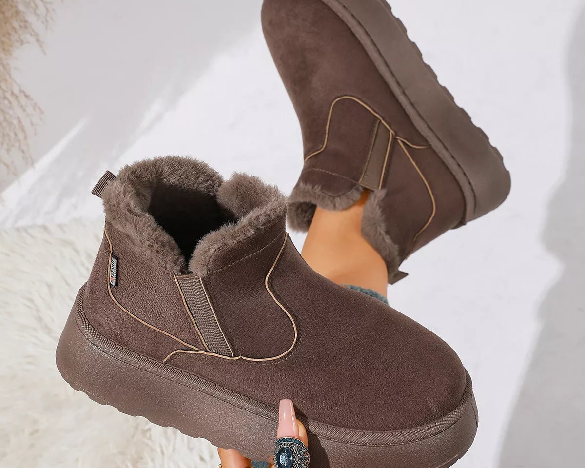 NEWYORKLISTED SHOE COLLECTION: A pair of women's mid-calf winter boots in dark brown suede with plush faux-fur lining, shown being worn with one boot raised and the other held by a hand. The boots feature elastic side panels and a thick lug sole.