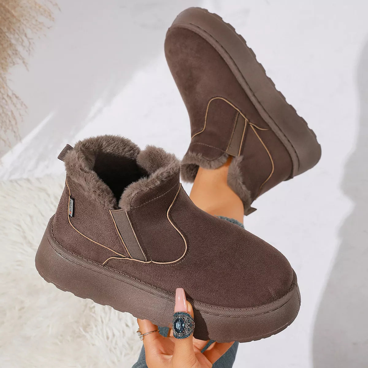 NEWYORKLISTED SHOE COLLECTION: A pair of women's mid-calf winter boots in dark brown suede with plush faux-fur lining, shown being worn with one boot raised and the other held by a hand. The boots feature elastic side panels and a thick lug sole.