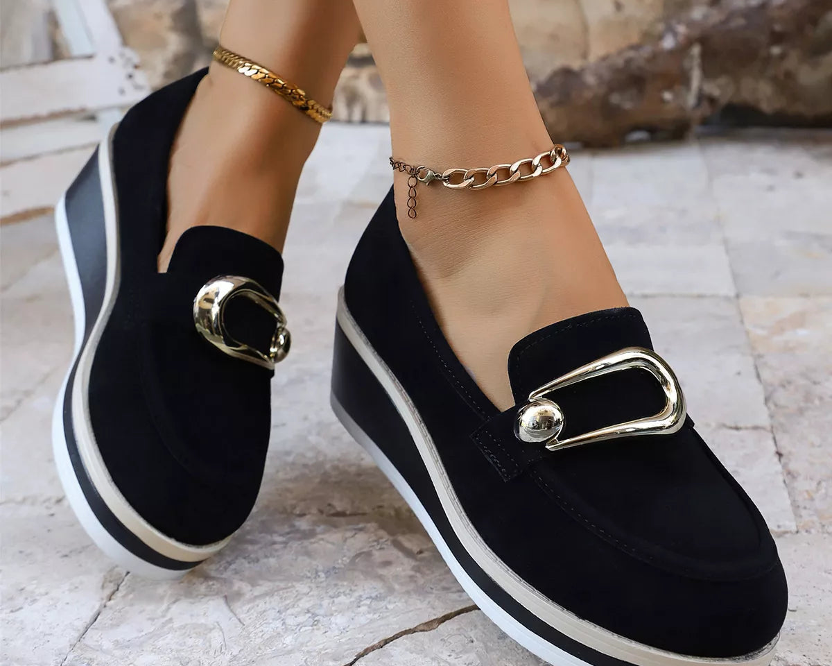 NEWYORKLISTED SHOE COLLECTION: Black loafers with gold buckles worn by a person on a light stone floor.