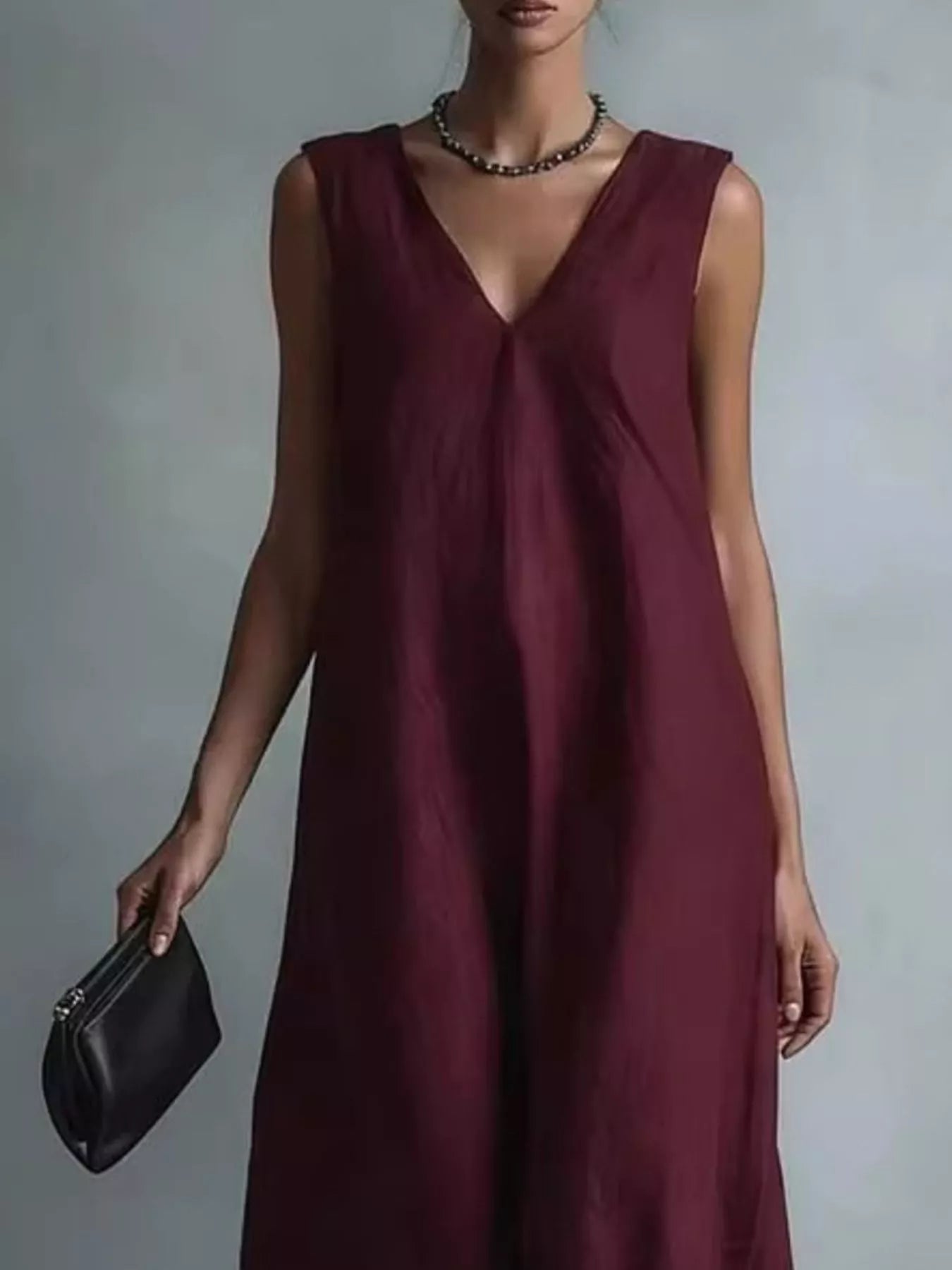 NEWYORKLISTED STYLE COLLECTION: A woman wearing a sleeveless deep burgundy V-neck maxi dress, holding a small black leather clutch against a neutral gray studio background.