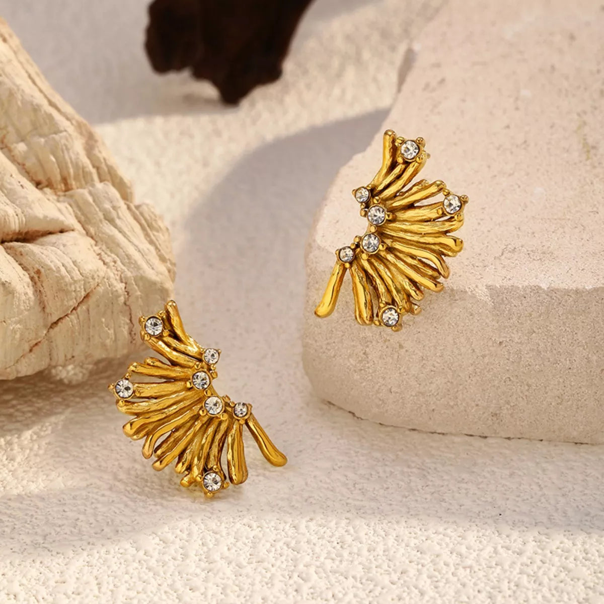 NYLD Glamour Collection by NEWYORKLISTED: Gold leaf-shaped earrings with rhinestones on a textured beige surface