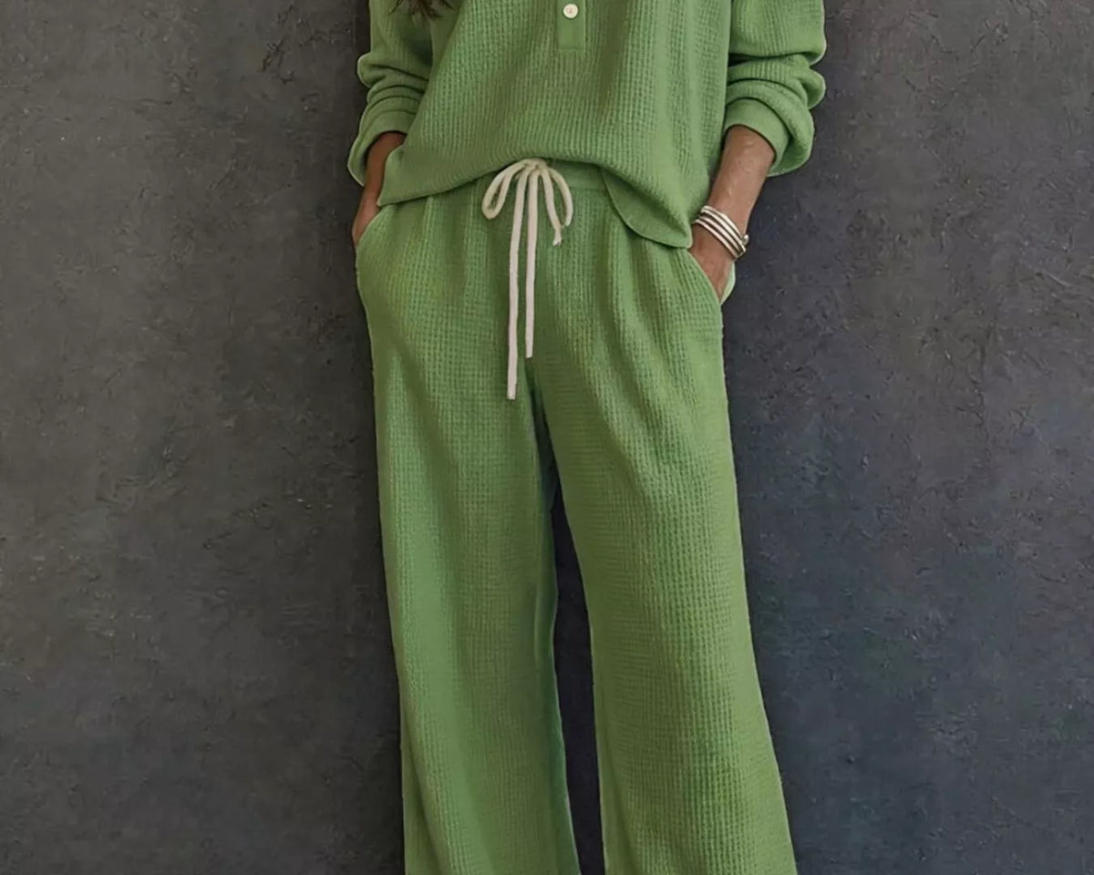 NEWYORKLISTED STYLE & COMFORT COLLECTION: A woman wearing a matching sage green knit lounge set consisting of a long-sleeve button-front top and wide-leg pants with a drawstring waist, standing against a dark textured wall.