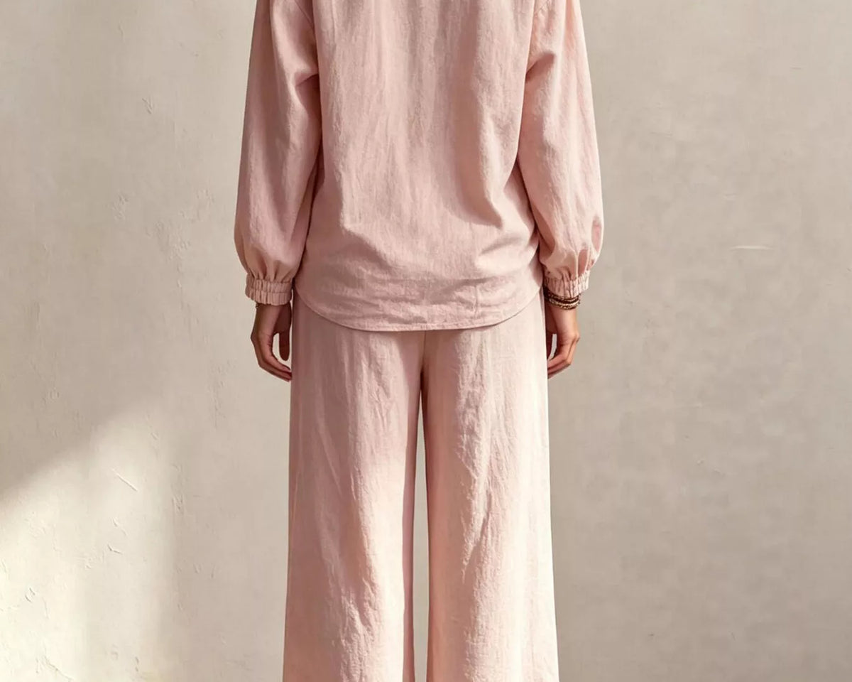 NEWYORKLISTED STYLE COLLECTION: A woman viewed from behind wearing a light pink linen two-piece set consisting of a long-sleeve top with gathered cuffs and matching wide-leg pants, standing against a textured beige wall.