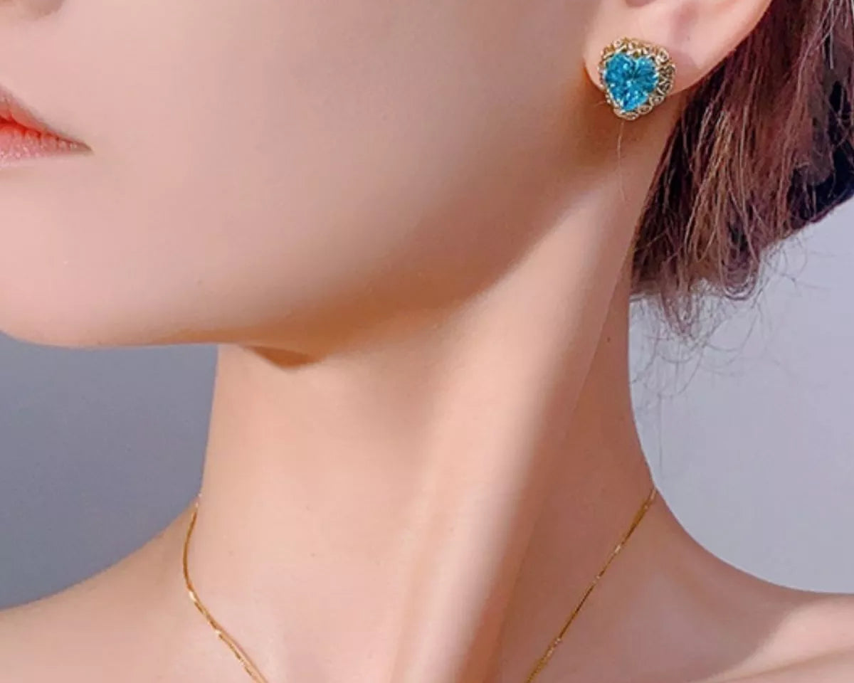 NYLD GLAMOUR COLLECTION By NEWYORKLISTED: Close-up of a woman wearing blue heart-shaped earrings with a blurred background