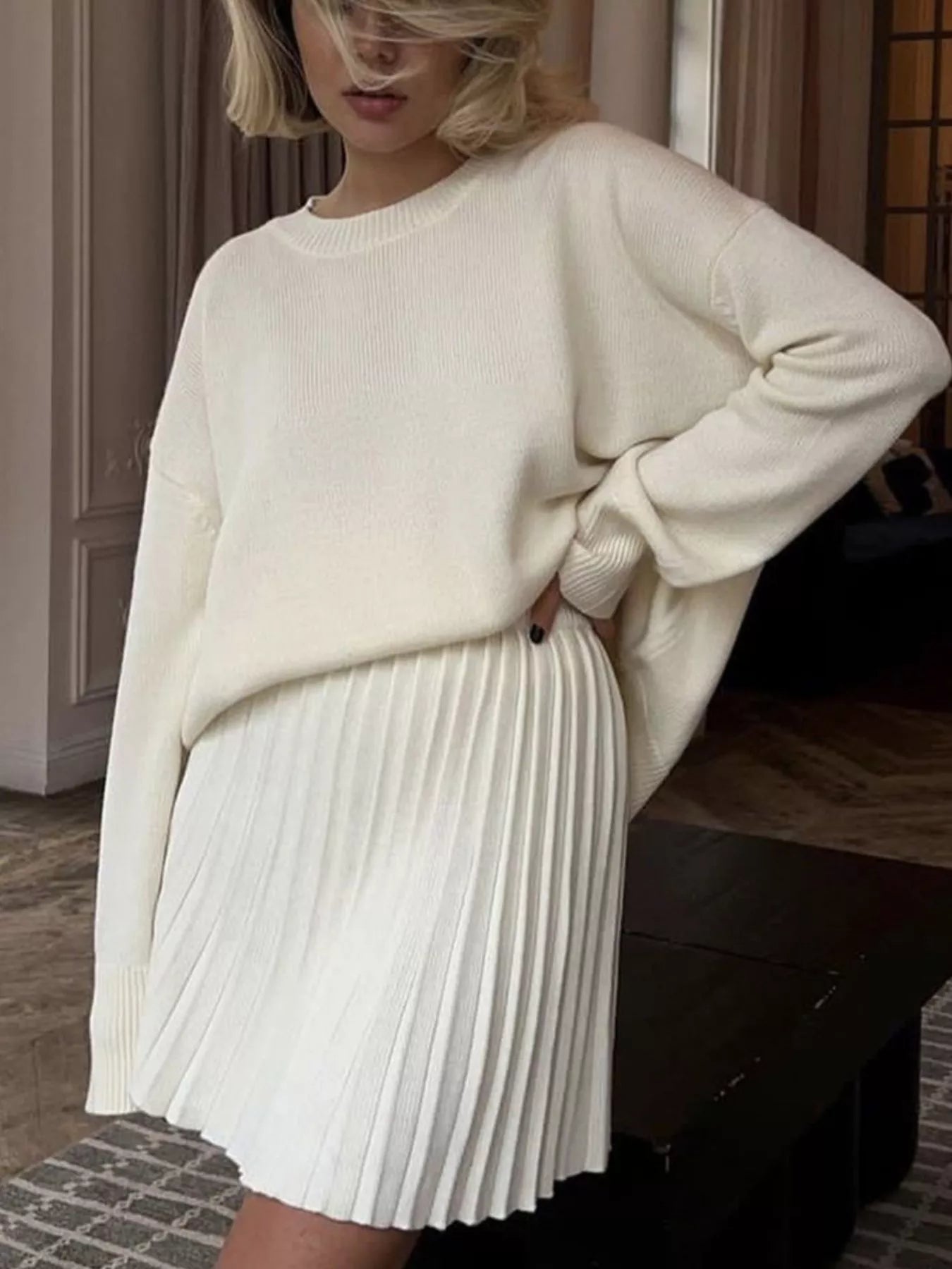 NEWYORKLISTED STYLE COLLECTION: Person wearing a cream sweater and white pleated skirt indoors.