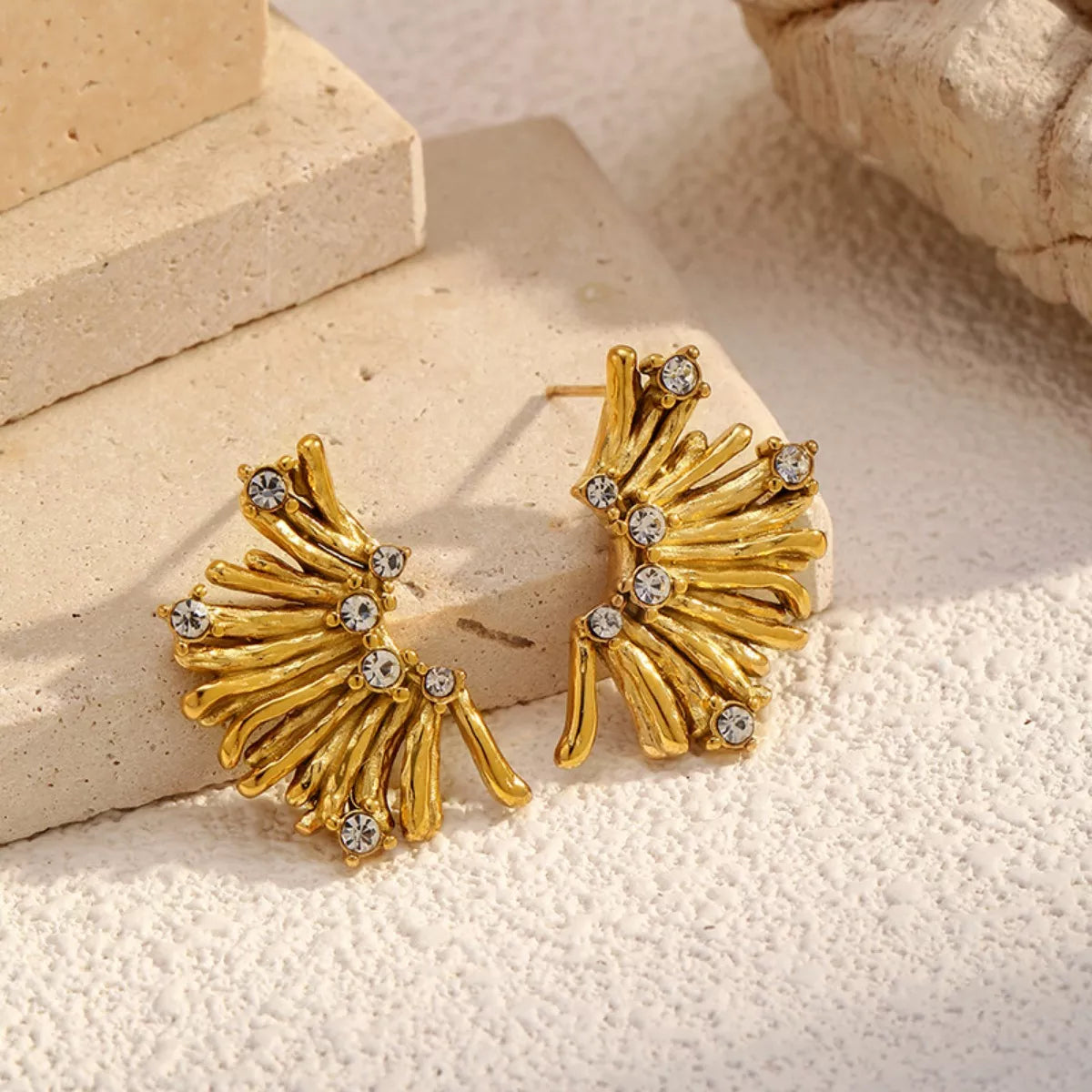 NYLD Glamour Collection by NEWYORKLISTED: Gold earrings with fan-like design and embedded stones on a textured surface