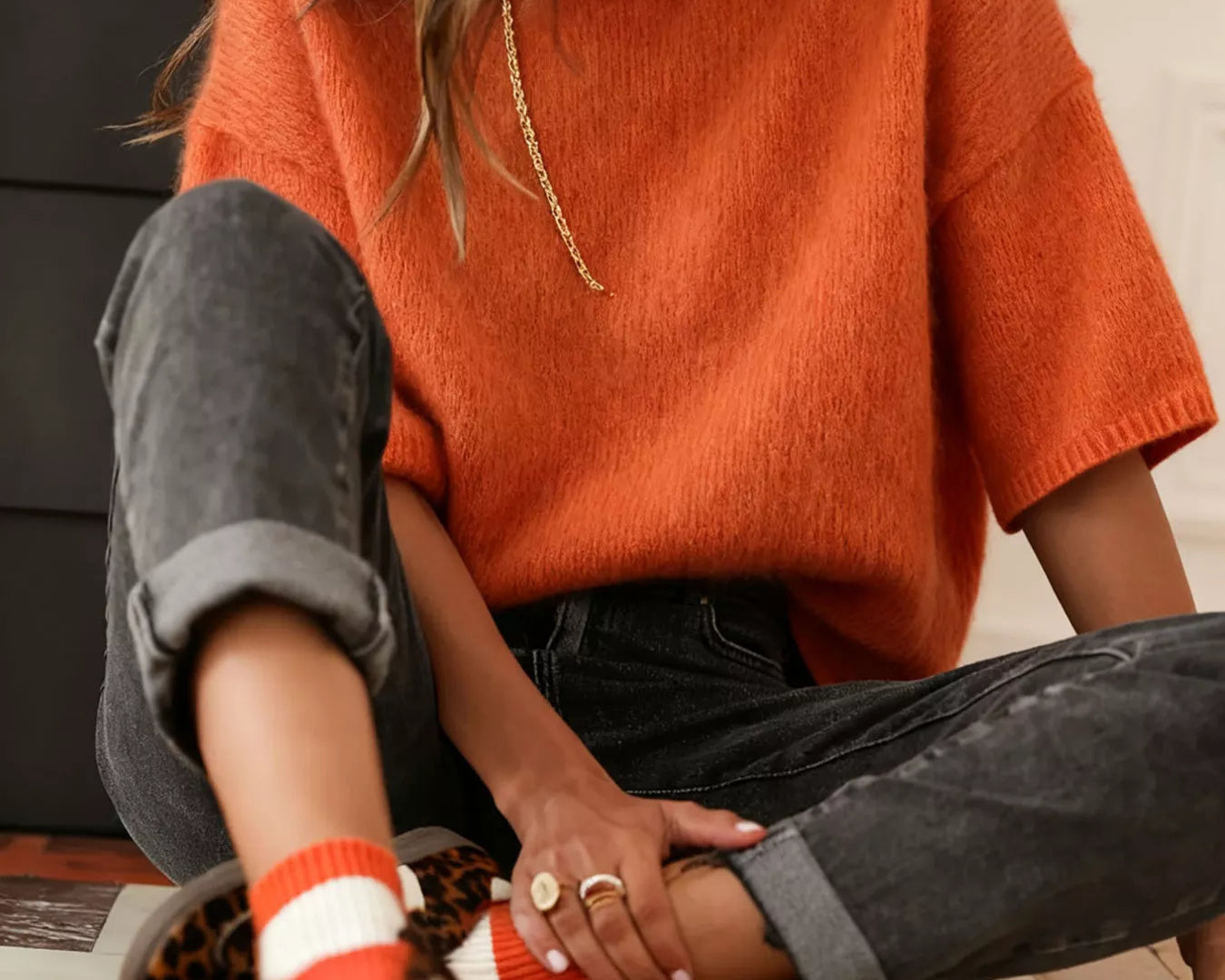 NEWYORKLISTED Style Collection: Person wearing an orange sweater and black pants sitting on a wooden floor.