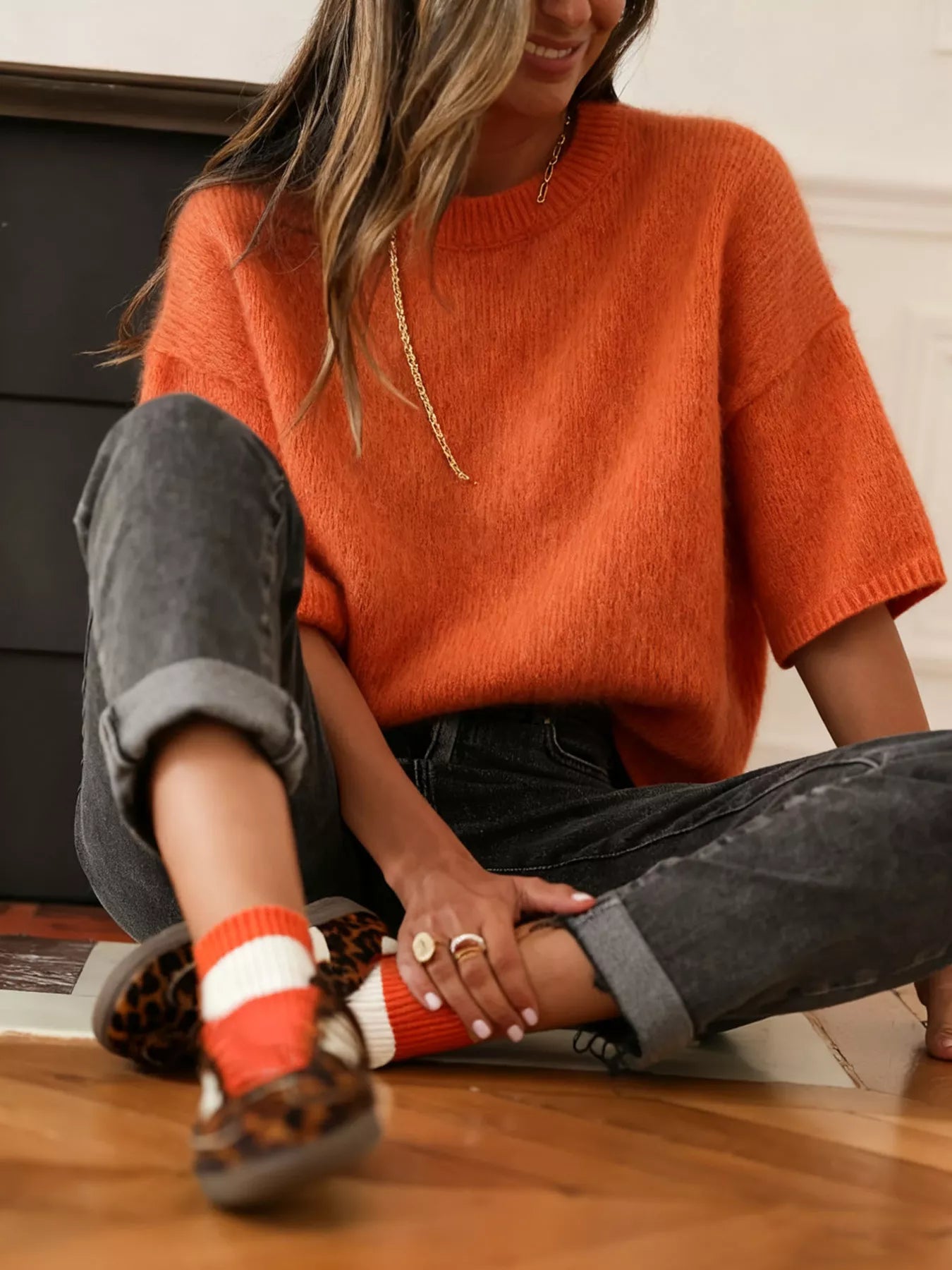 NEWYORKLISTED Style Collection: Person wearing an orange sweater and black pants sitting on a wooden floor.