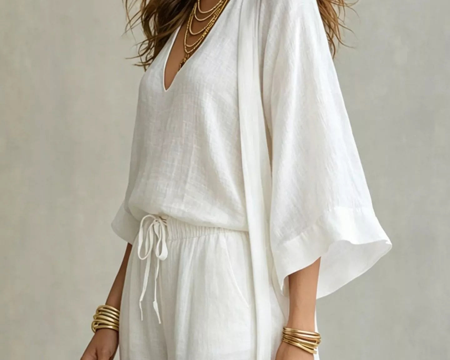 NEWYORKLISTED STYLE COLLECTION: A woman wearing a white linen kaftan-style dress with wide bell sleeves, a V-neckline, and a drawstring waist, styled with layered gold necklaces, bracelets, hoop earrings, and a woven straw handbag against a neutral textured background.