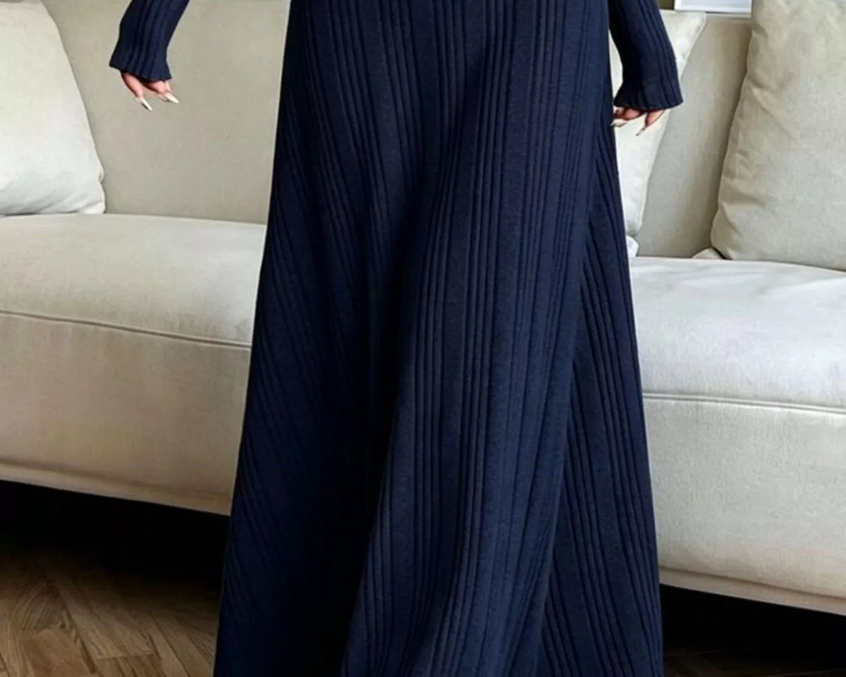 NEWYORKLISTED STYLE COLLECTION: A person wearing a long-sleeve, floor-length navy blue ribbed knit maxi dress, standing in a living room with a beige sofa and wood floor.