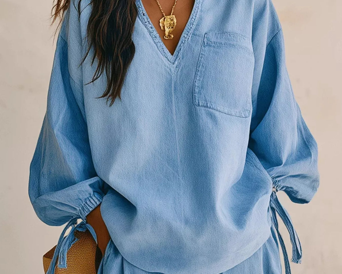 NEWYORKLISTED STYLE COLLECTION: A woman wearing a light blue oversized long-sleeve top with a V-neck, chest pocket, and side tie details, paired with matching light blue bottoms. She is accessorized with gold jewelry and blue earrings against a neutral beige background.