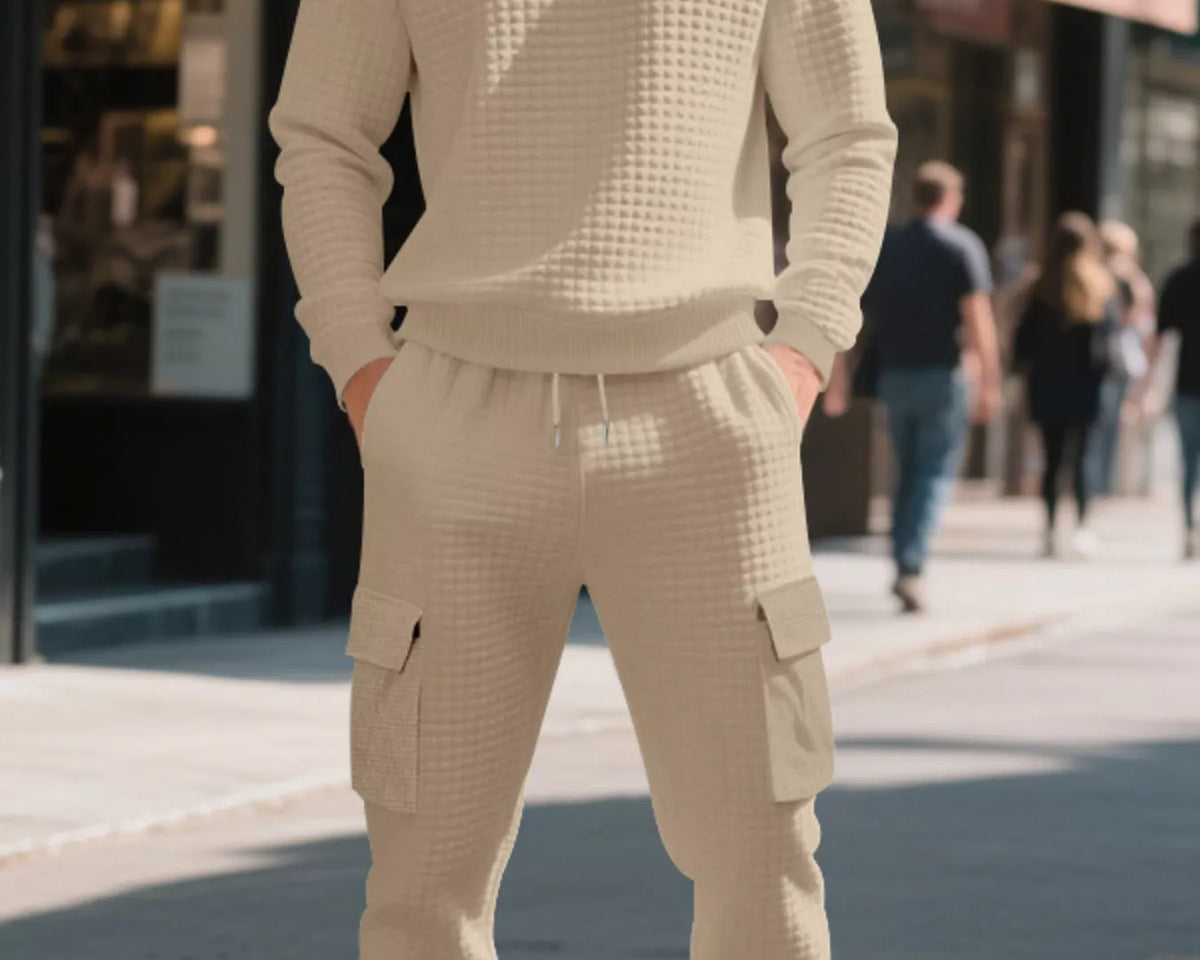 NEWYORKLISTED ATLAS MEN'S COLLECTION: A man wearing a matching beige waffle-knit sweatshirt and cargo jogger pants stands on a city sidewalk, with blurred storefronts and pedestrians in the background.