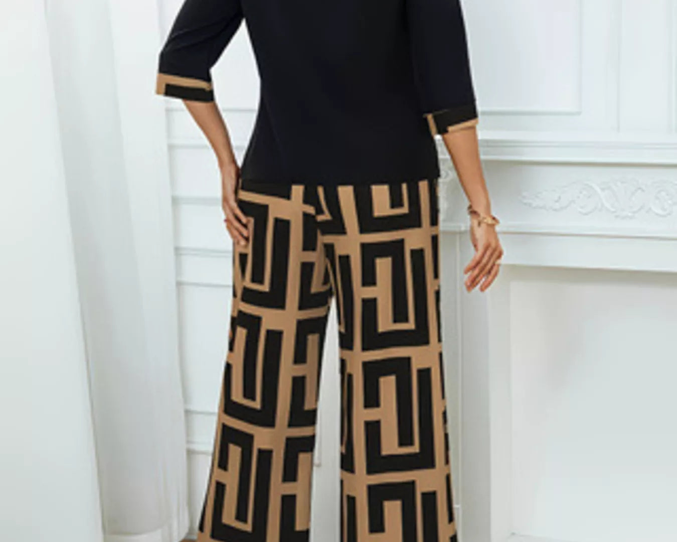 NEWYORKLISTED: Woman wearing a black top and patterned pants standing in a room with white curtains.