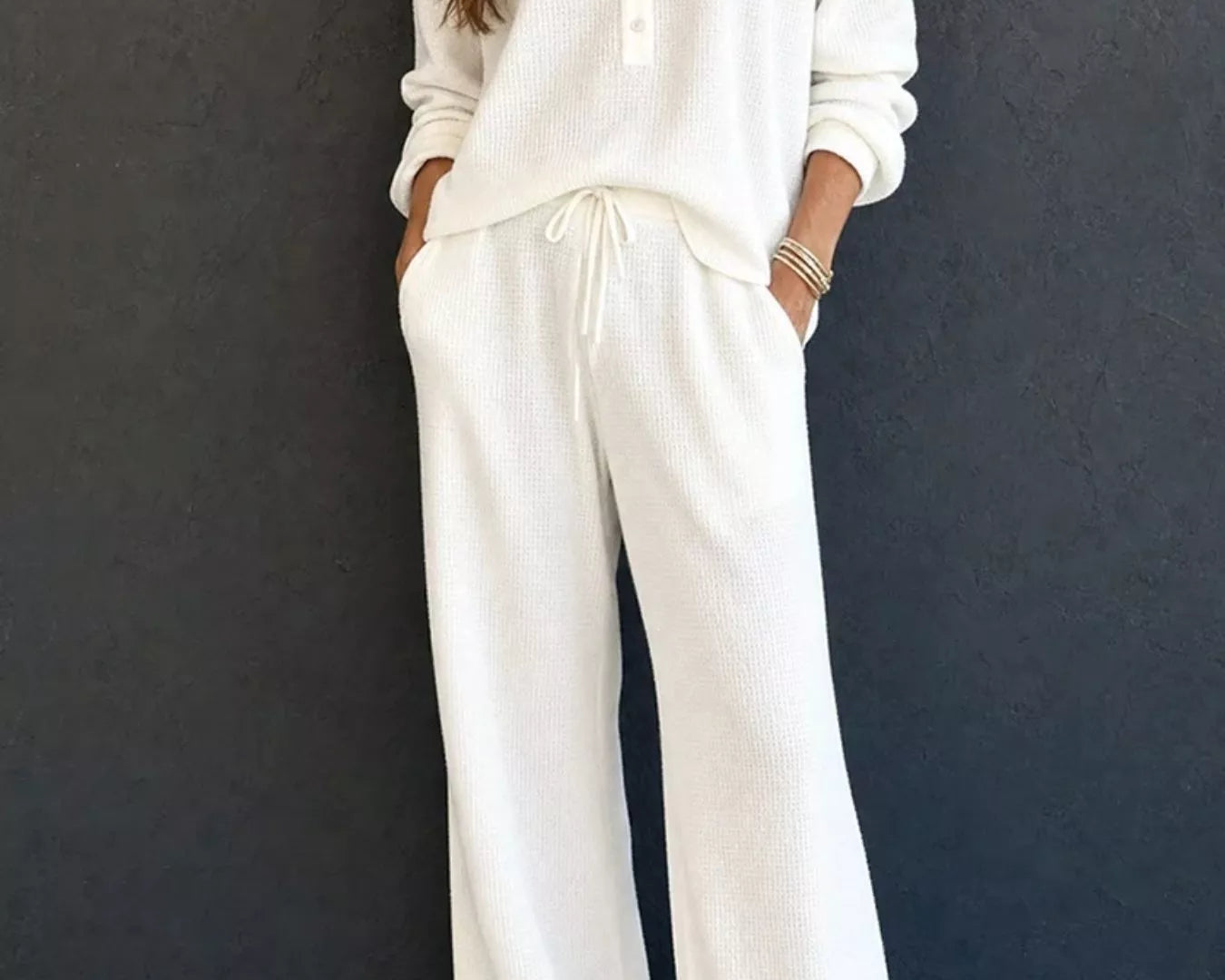 NEWYORKLISTED STYLE & COMFORT COLLECTION: A woman wearing a matching off-white knit lounge set consisting of a long-sleeve henley top and wide-leg drawstring pants, standing against a dark textured wall.