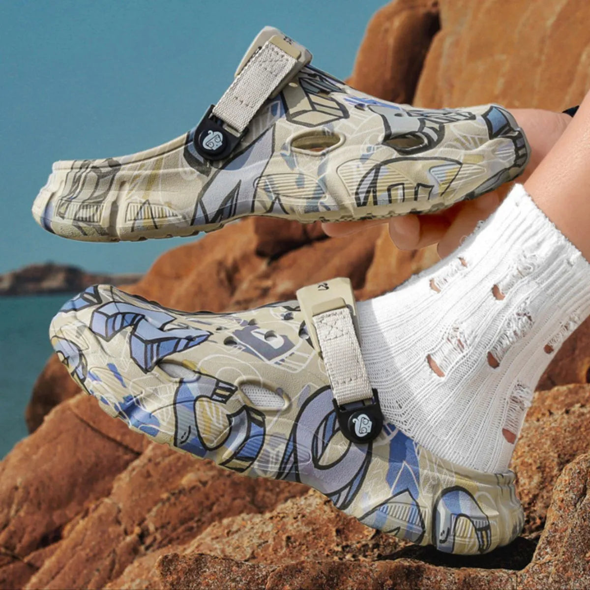 NEWYORKLISTED ATLAS MEN'S COLLECTION: Pair of patterned sandals held by a person on rocky terrain with water in the background