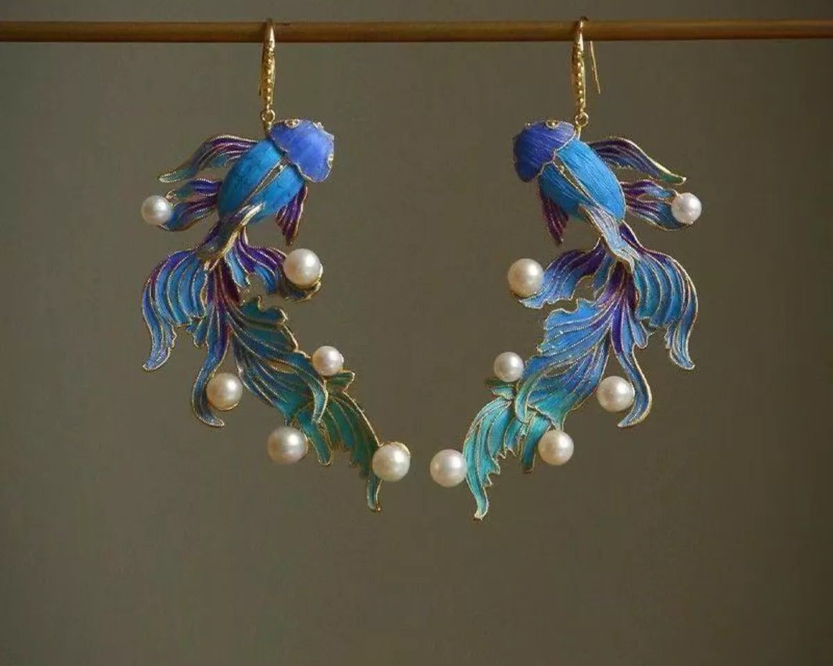 NYLD GLAMOUR By NEWYORKLISTED: Pair of blue fish-shaped earrings with pearl accents on a dark background