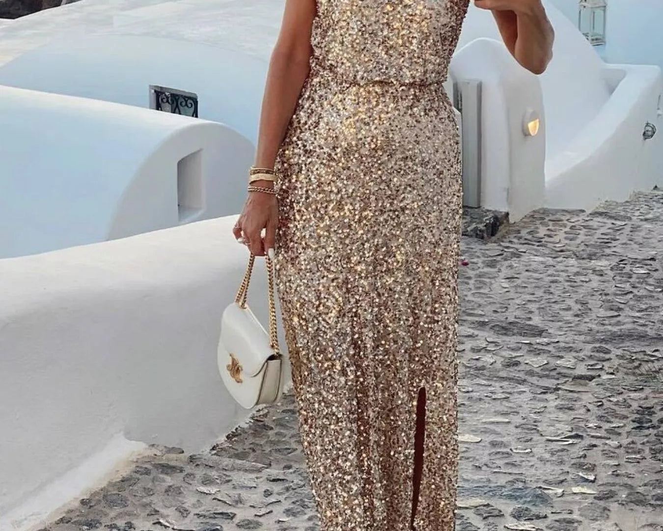 NEWYORKLISTED STYLE COLLECTION: Woman in a sparkly one-shoulder dress standing on a cobbled street with white buildings and blue sea in the background.
