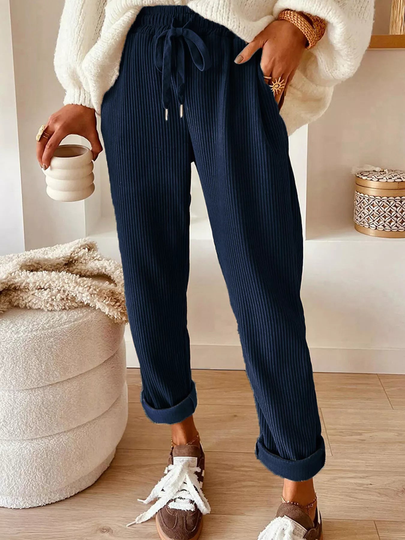 NEWYORKLISTED STYLE COLLECTION: Person wearing navy ribbed pants with a white sweater and brown shoes, holding a small white container.