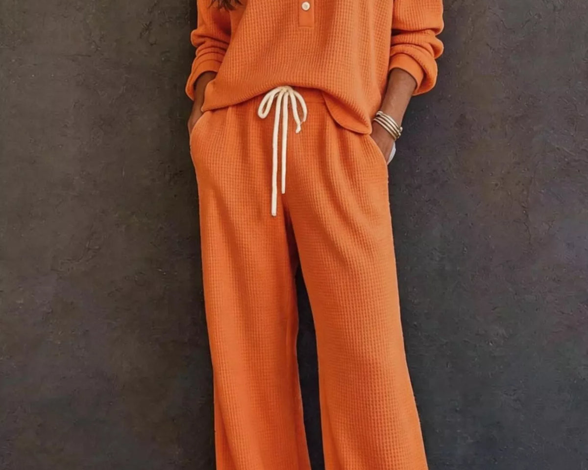 NEWYORKLISTED STYLE & COMFORT COLLECTION: A woman wearing a matching orange waffle-knit lounge set with a long-sleeve henley top and wide-leg pants, featuring a white drawstring, standing against a dark textured wall.