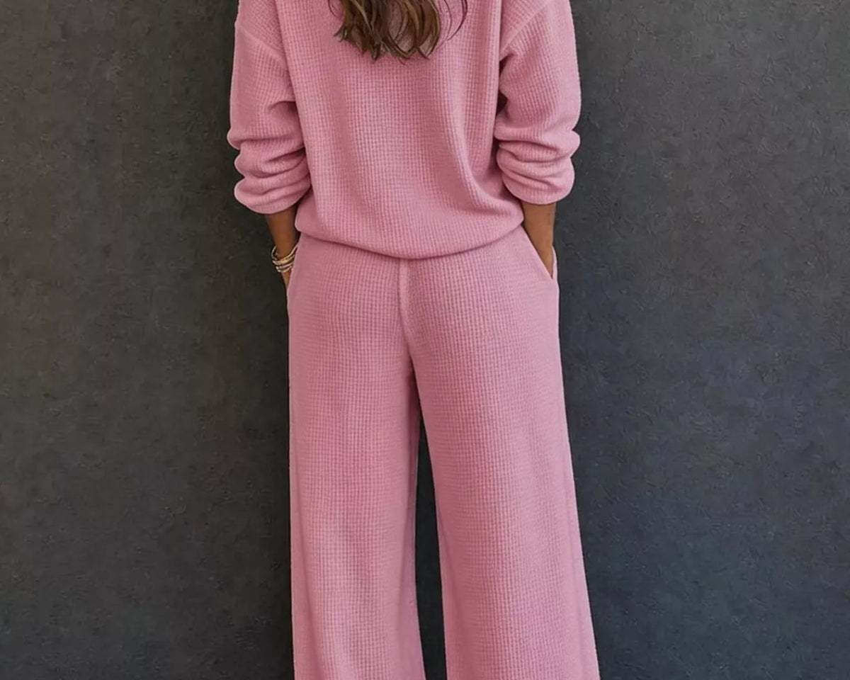 NEWYORKLISTED STYLE & COMFORT COLLECTION: A woman viewed from behind wearing a matching dusty pink knit lounge set consisting of a long-sleeve top and wide-leg pants, standing against a dark gray textured wall.