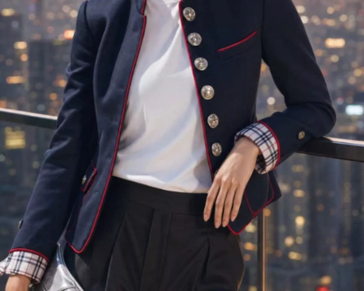 NEWYORKLISTED STYLE COLLECTION: A woman models a navy blue blazer with red piping and silver buttons, worn over a white top and black trousers, set against a city skyline at sunset.