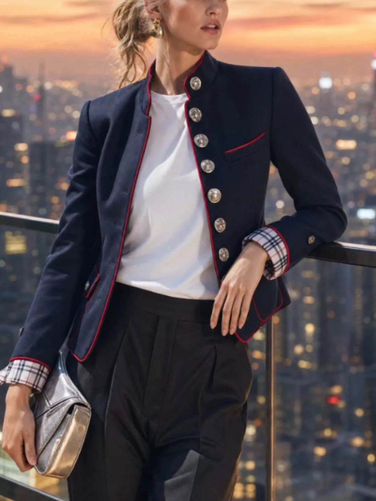 NEWYORKLISTED STYLE COLLECTION: A woman models a navy blue blazer with red piping and silver buttons, worn over a white top and black trousers, set against a city skyline at sunset.