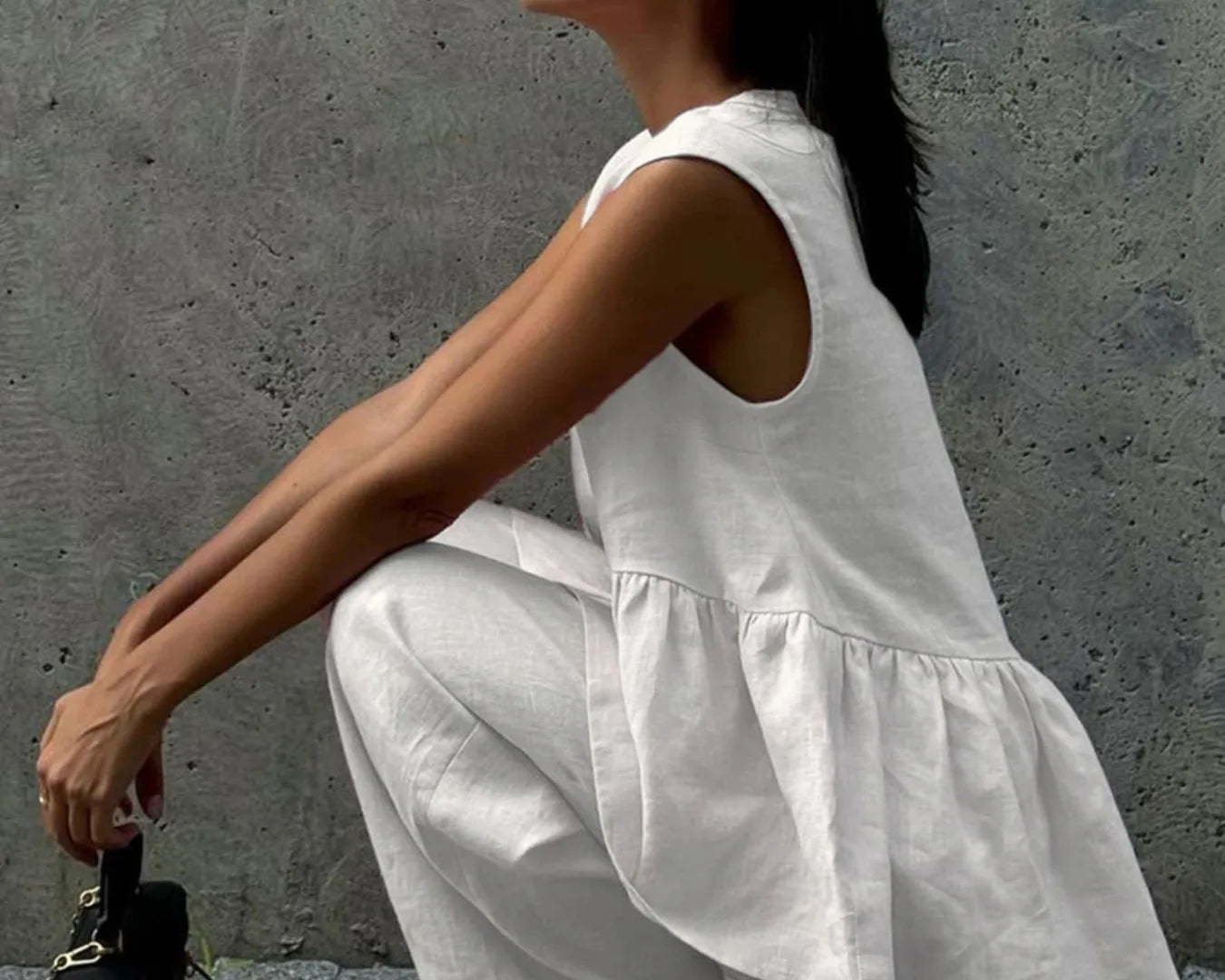 NEWYORKLISTED STYLE COLLECTION: Woman in a white sleeveless top and pants squatting against a gray wall.
