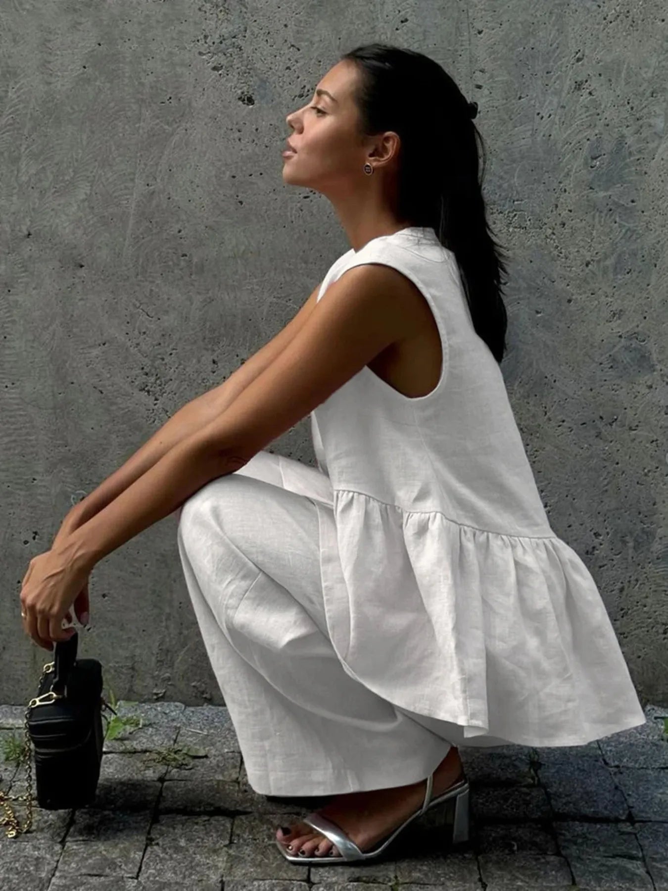 NEWYORKLISTED STYLE COLLECTION: Woman in a white sleeveless top and pants squatting against a gray wall.