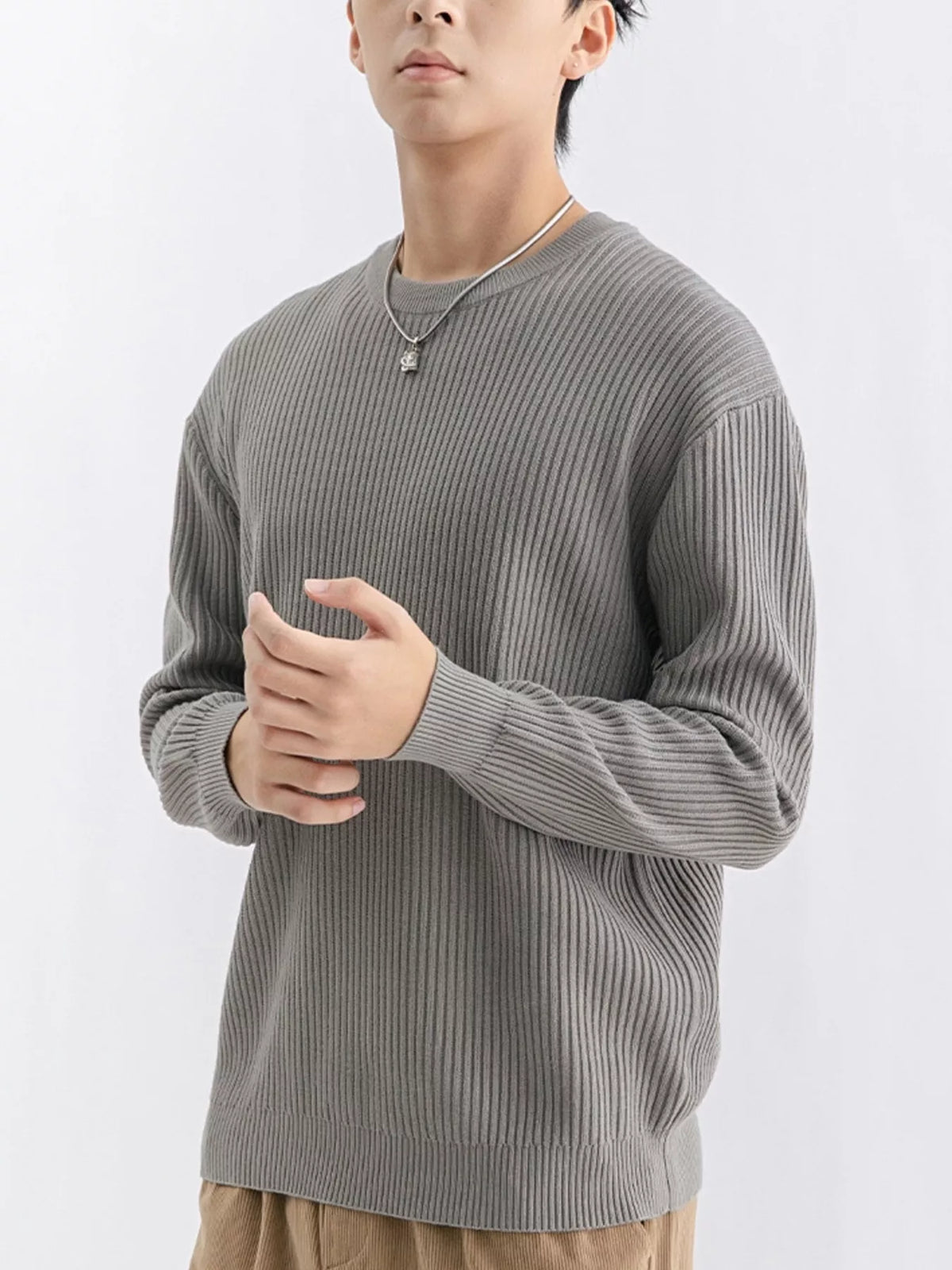 NEWYORKLISTED ATLAS MEN'S COLLECTION: A man wearing a ribbed gray long-sleeve crewneck sweater, paired with tan pants and a silver necklace, photographed against a clean white studio background.