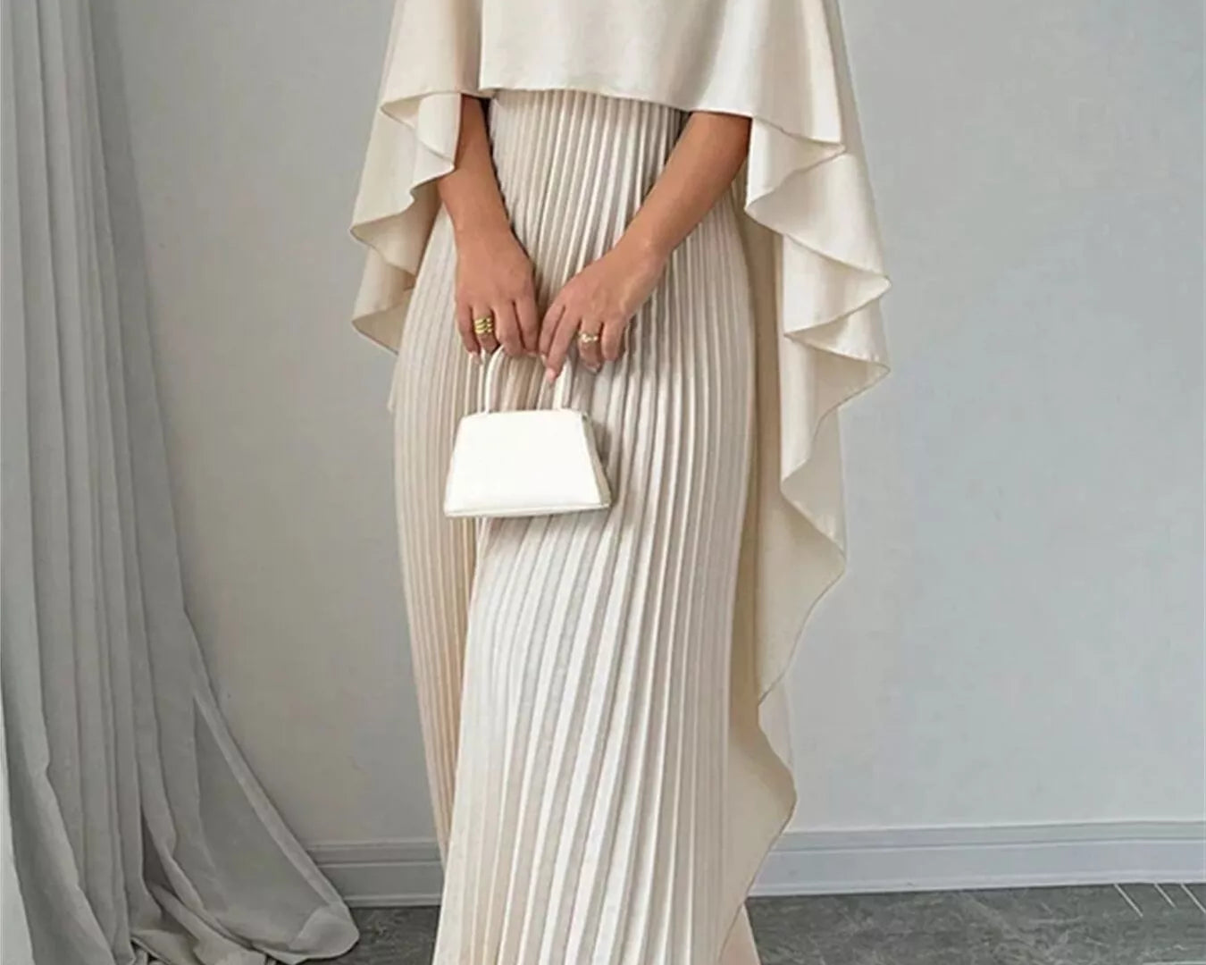 NEWYORKLISTED STYLE COLLECTION: Woman wearing a beige cape over a pleated dress holding a white clutch against a plain background