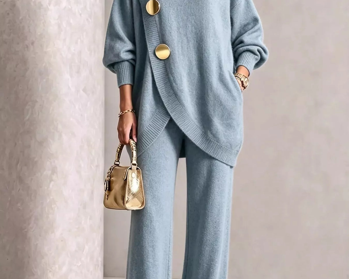 NEWYORKLISTED STYLE COLLECTION: A woman models a light blue knit two-piece set consisting of an asymmetrical turtleneck sweater with large gold buttons and matching wide-leg trousers. She is holding a small metallic gold handbag and wearing gold pointed shoes, set against a neutral textured beige background.
