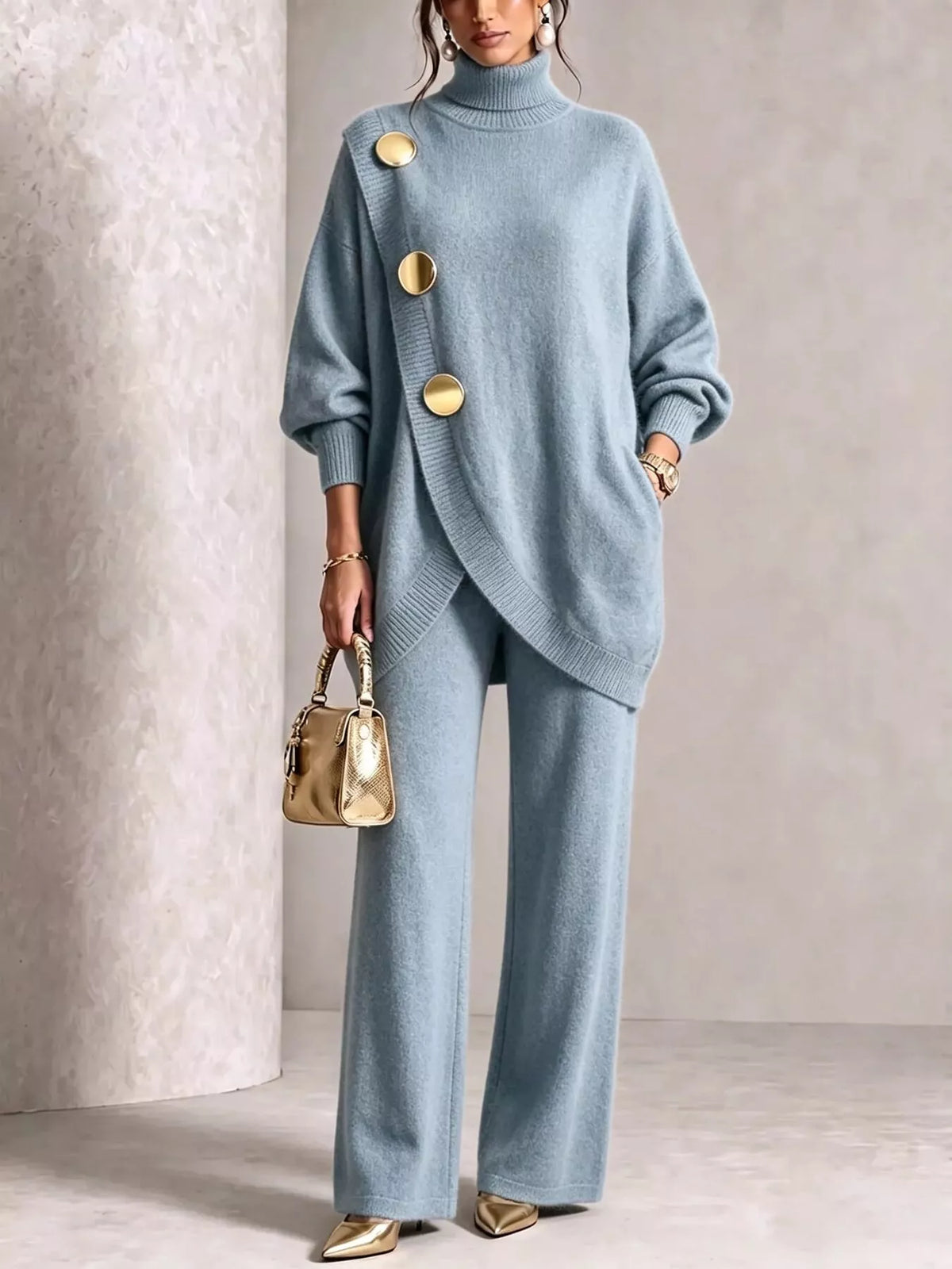 NEWYORKLISTED STYLE COLLECTION: A woman models a light blue knit two-piece set consisting of an asymmetrical turtleneck sweater with large gold buttons and matching wide-leg trousers. She is holding a small metallic gold handbag and wearing gold pointed shoes, set against a neutral textured beige background.