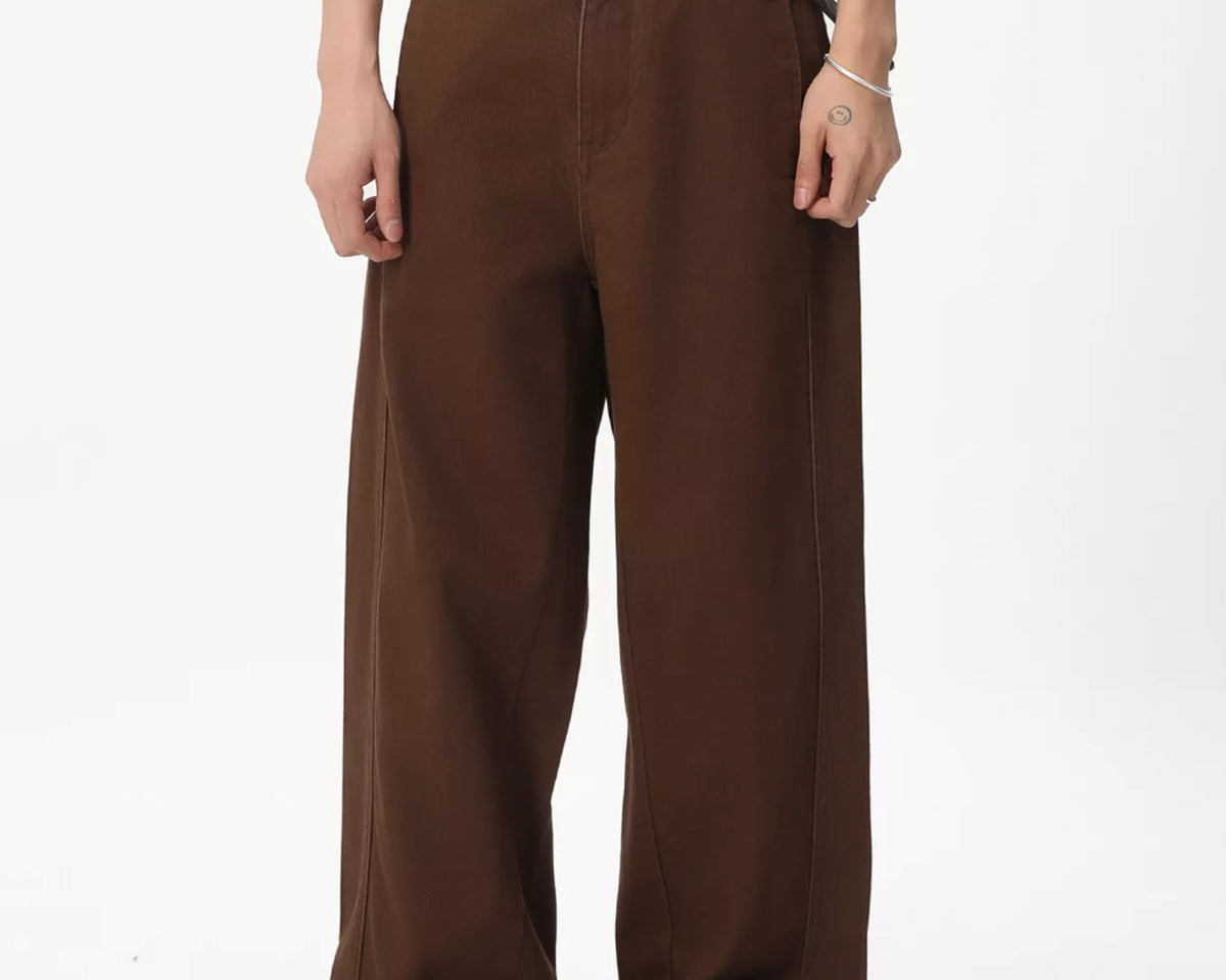 NEWYORKLISTED ATLAS MEN'S COLLECTION: A person wearing loose brown wide-leg pants with a dark belt and a small red heart-shaped buckle detail, paired with a white graphic T-shirt and tan shoes, photographed against a plain white studio background.